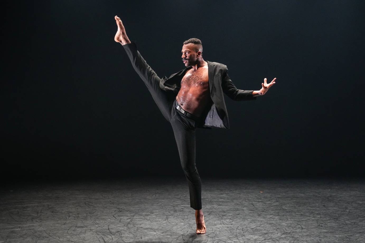 a Black man in tight black pants, and an open black jacket, his chest is bare, kicks one leg very high to his side, he has an intent concentrated look on his face. The kick is impressive.